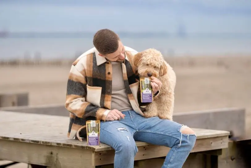 Hund und Halter sitzen auf einem Steg am Strand, der Hund schnüffelt an den Nassfutter Pouches von Wildes Land.
