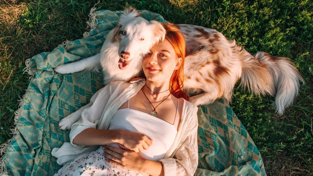 Eine junge Frau mit roten Haaren liegt mit einem lächelnden braun-weiß gefleckten Hund auf einer Decke im Gras. Beide genießen die Sonne in einem entspannten Moment im Freien.