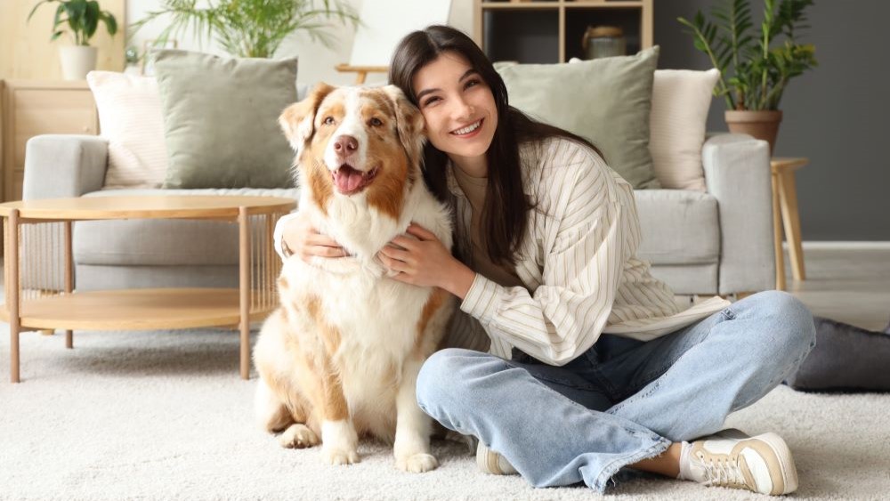 Eine lächelnde Frau sitzt in einem gemütlichen Wohnzimmer auf dem Teppich neben einem braun-weißen Hund, den sie umarmt. Im Hintergrund sind Sofa, Pflanzen und Regale zu sehen.