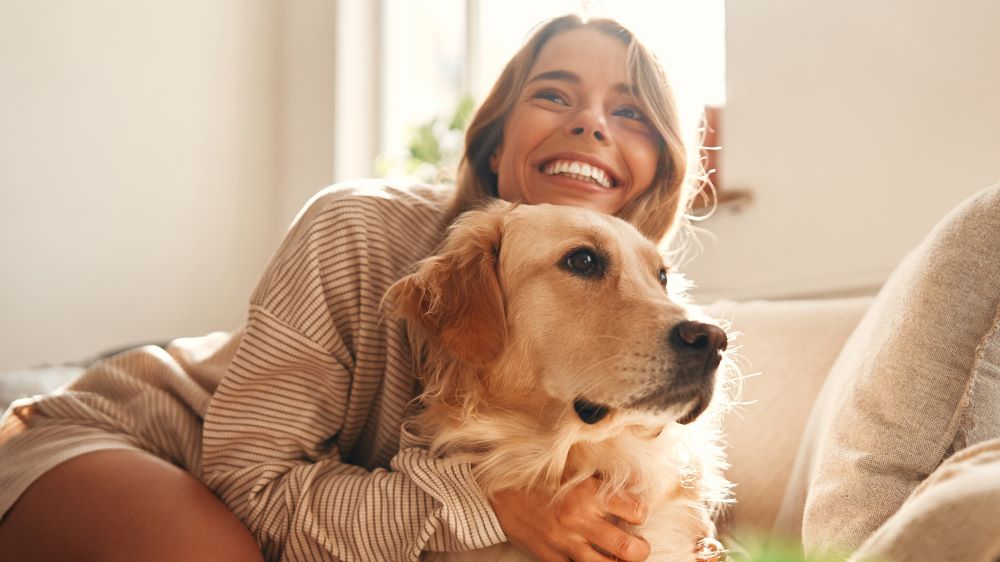Eine strahlende Frau kuschelt mit einem Golden Retriever auf einem Sofa in einem sonnigen Raum. Beide wirken glücklich und entspannt.