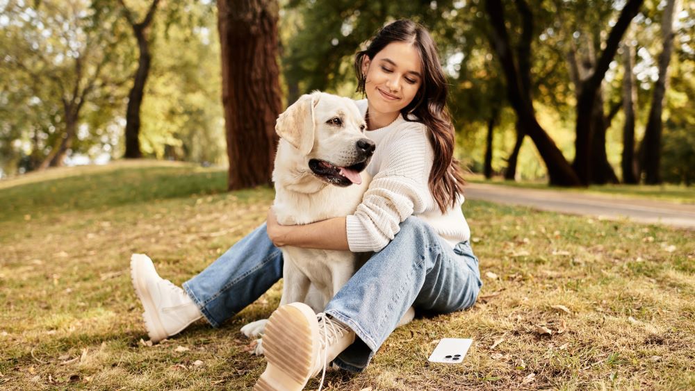 Eine Frau sitzt auf dem Rasen in einem Park und umarmt einen Labrador, beide wirken zufrieden.