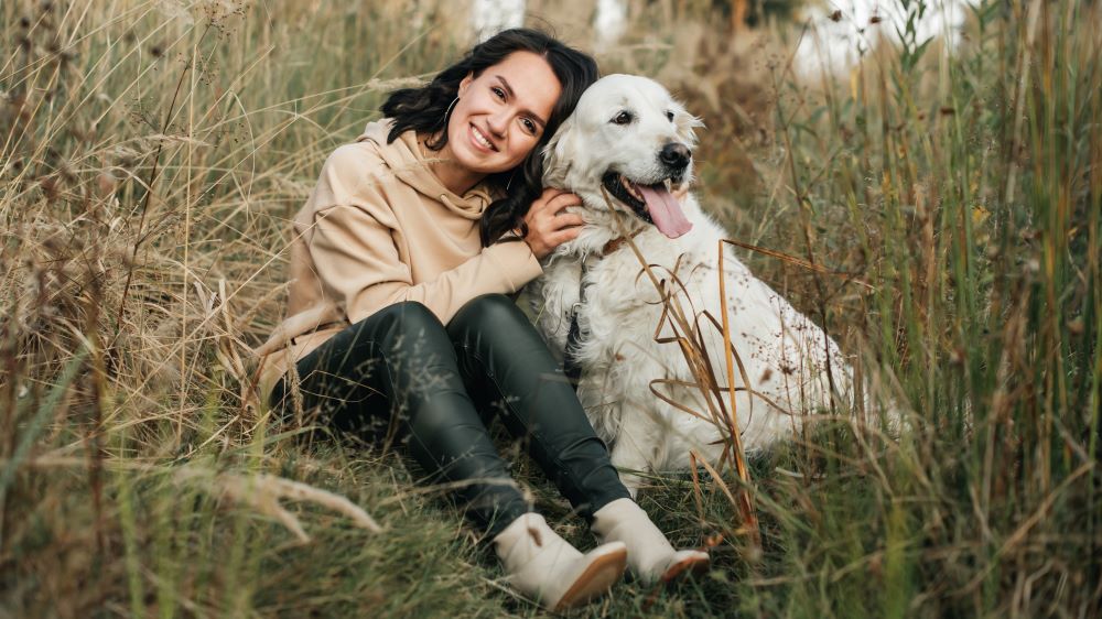 Eine lächelnde Frau sitzt in hohem Gras und kuschelt mit einem großen, weißen Hund. Beide wirken entspannt und genießen die Natur.