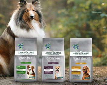 A long-haired collie sits outdoors behind three bags of Arden Grange Grain Free dry dog food, each featuring different recipes and dog images on the packaging.